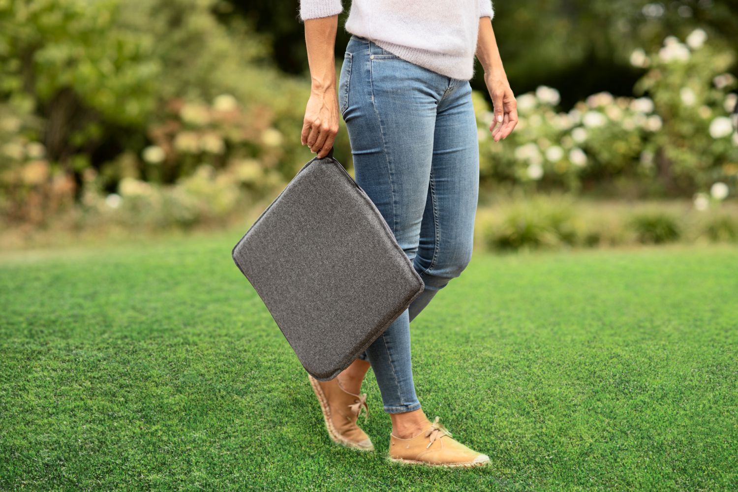 A person carries a gray padded seat cushion while walking across a green lawn.